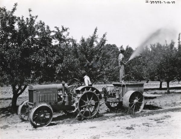 A man drives a McCormick-Deering 10-20(?) tractor through an orchard as another man stands on an orchard sprayer while applying what is probably pesticide to the trees.