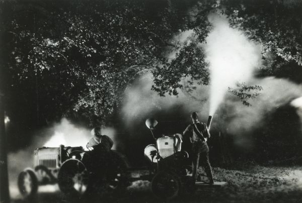 Two men use a McCormick-Deering tractor and an orchard sprayer to apply pesticide to trees at night.