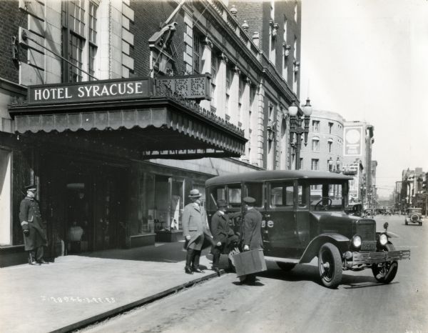 A bellhop is holding a suitcase while holding the door of an International Model S speed sedan open for guests at Hotel Syracuse. The truck is parked under the front awning of the hotel, and other commercial buildings line both sides of the street in the background.