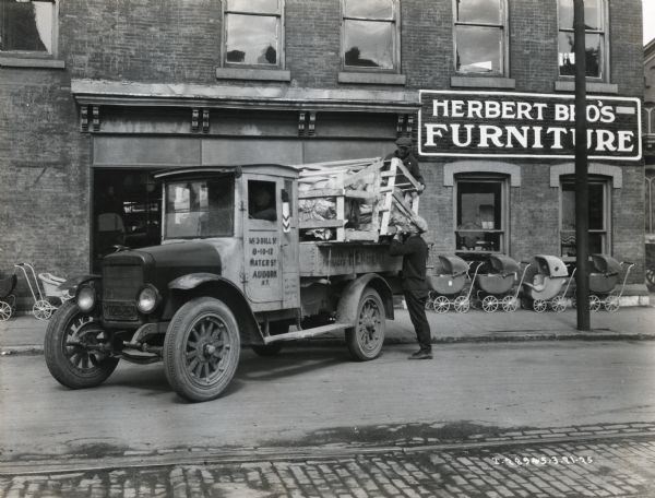 A man is loading wooden crates onto the bed of an International Model S  truck owned by Herbert Brothers furniture merchants. A row of baby strollers is lined up in front of the storefront behind the truck.
