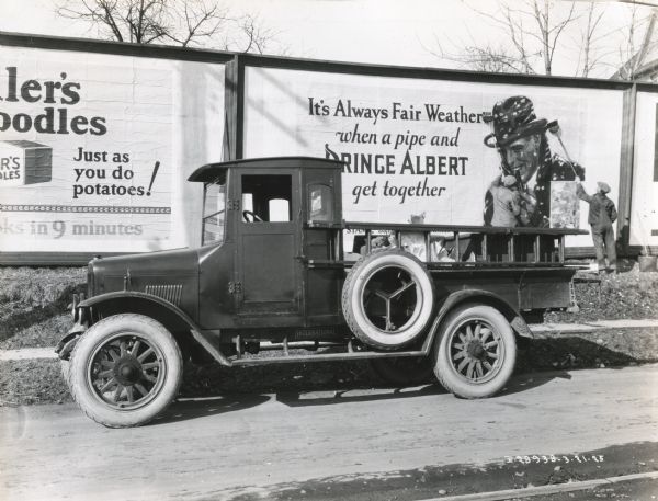 International Model S truck owned by the Wallace Poster Advertising Company is parked along the side of a road in front of two billboards. Two men on the right are using brooms to affix a "Prince Albert" poster onto the billboard.