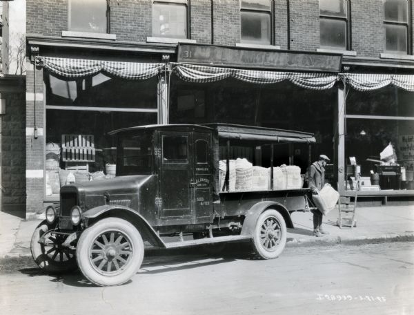 A man loads a burlap bag of seeds onto the back of an International Model S truck owned by D.L. Ramsey & Son. The truck is parked along the curbside in front of the Ramsey storefront.