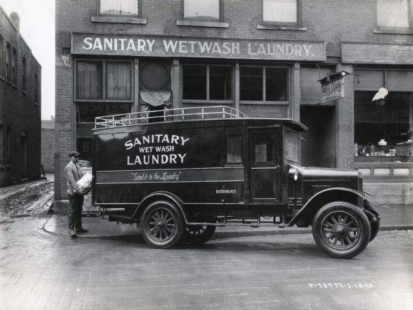 A man loads a bag of laundry onto the back of an International Model S truck owned by the Sanitary Wet Wash Laundry. The truck is parked in front of the company's storefront and the text on the truck reads, "Send it to the Laundry."