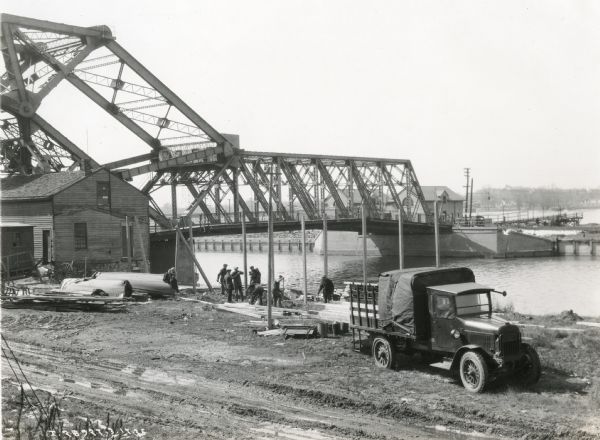 An International Model 43 truck owned by Whitmier & Ferris Company parked along the bank of a river where men are constructing the framework to a billboard. A bridge spans the water in the background.
