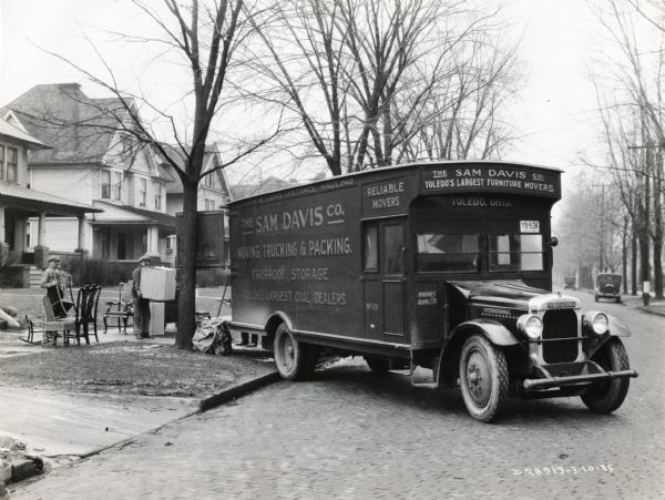 Furniture movers from the Sam Davis Company load the back of a Model 53 International motor truck with chairs and boxes. The text on the truck reads: "The Sam Davis Co. Moving, Trucking, & Packing. Fireproof Storage. Toledo's Largest Coal Dealers."