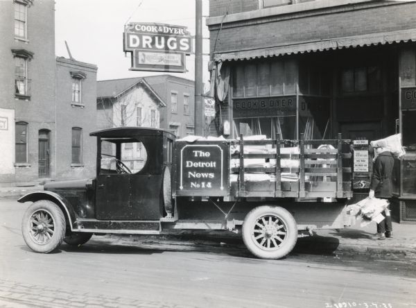 An International Model SL truck owned by "The Detroit News" parked in front of Cook & Dyer Drugstore. A man is removing piles of newspapers from the truck bed during a delivery to the store.