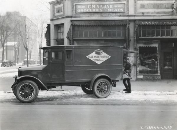 An International Model S truck owned by the Blue Valley Creamery Company is parked outside a grocery store as a man unloads goods from the enclosed truck bed.