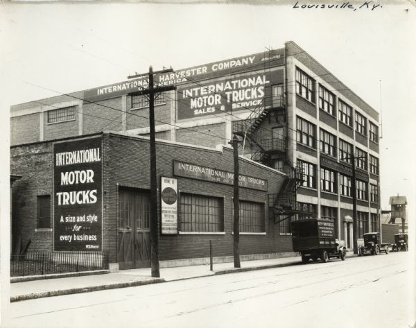 Exterior view of the International Harvester Company Louisville branch. Several automobiles are parked along the street in front of the building. On the far right is an elevated guardhouse or shed near a brick wall.