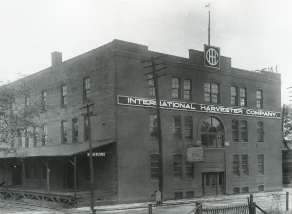 Exterior view of the International Harvester Company Knoxville branch.  A sign advertising P&O plows hangs from a covered porch along the side of the building and the IHC logo appears along the roof. A mailbox stands beneath a power line beside the building.