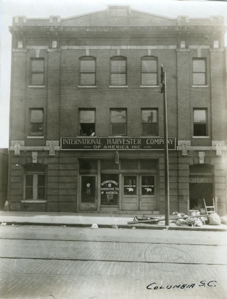 A is man looking out the window of the second story of the International Harvester Company's Columbia branch building. A sign is hanging above the main entrance, and an open elevator shaft is on the right side of the building's facade. Bundles and a handcart are on the sidewalk nearby. A datestone at the top of the building reads: "1810."