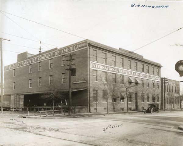 External view of International Harvester Company's Birmingham branch building, located at the intersection of 10th Avenue and 24th Street.  A railroad car stands beside the building. Railroad tracks are across the street in the foreground. An automobile is parked along the curb on the right.