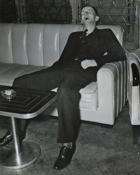 An International Harvester dealer, T.F. Hogan, slouches in a couch and sleeps at a convention or meeting.