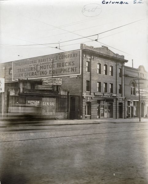 Exterior view of the International Harvester Company Columbia branch building. Brick buildings stand on either side; to the left is a building bearing the sign "General Blacksmith" and to the right is Colman-Drew Co. Wholesale Candy & Drugs."