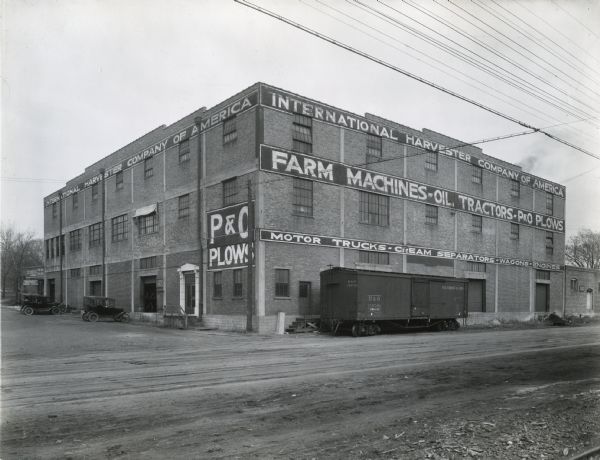 Exterior view of International Harvester's Springfield branch building. A railroad car stands beside the building and several automobiles are parked nearby. The sign painted on the building's brickwork reads, "International Harvester Company of America. Farm Machines - Oil Tractors - P&O Plows - Motor Trucks - Cream Separators - Wagons - Engines."