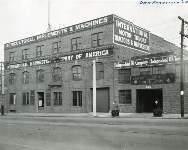 Exterior view of International Harvester's San Francisco branch. The brick building is attached to an adjacent building which bears signs reading: "Independent Oil Company" and "Hendrie Rubber Tire Co. 'Hendrie Cords.'" There are two wheeled carts sitting on the sidewalk, perhaps for pumps to inflate tires.