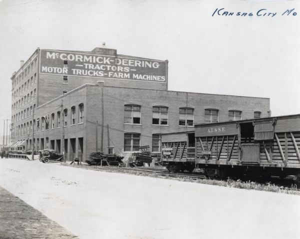 Train cars rest on tracks leading past International Harvester's Kansas City branch. Farm equipment stands on loading dock on the side of the building, and several automobiles are parked nearby.