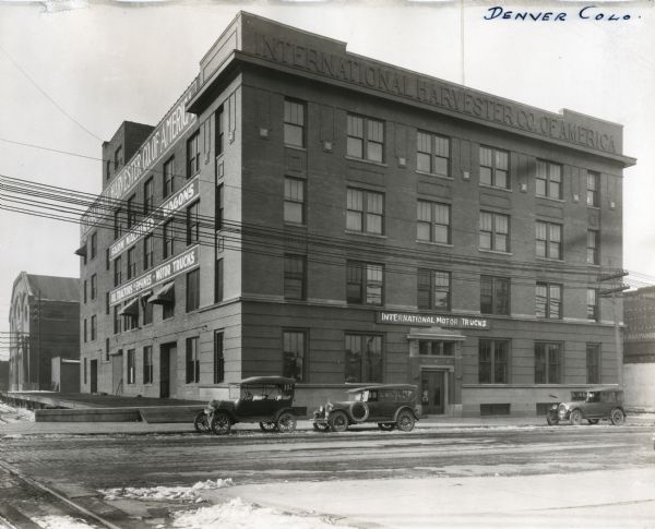 Exterior view of International Harvester Company's Denver branch office. Several automobiles park along the road in front of the building and signs on the side read, "Farm Machines - Wagons - Oil Tractors & Engines - Motor Trucks."