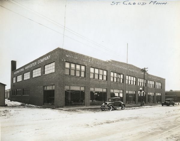 Automobiles are parked along the curb in front of International Harvester Company's St. Cloud branch. The signs on the building read, "International Harvester Company" and "Motor Trucks, Tractors, Farm Operating Equipment."
