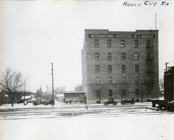 Exterior view from across street of the International Harvester Mason City branch building. Several automobiles and a horse-drawn wagon are parked outside; railroad tracks are in the foreground, and a railroad car is on the right near a raised platform.