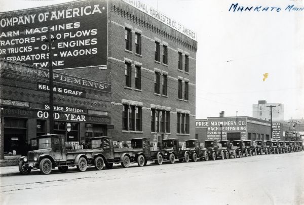 Exterior view of International Harvester's Mankato branch building with a long line of at least 17 trucks parked along the curb. The sign on the side of the building advertises: "Company of America. Farm Machines. Tractors - P&O Plows. Motor Trucks - Oil Engines. Cream Separators - Wagons. Twine."