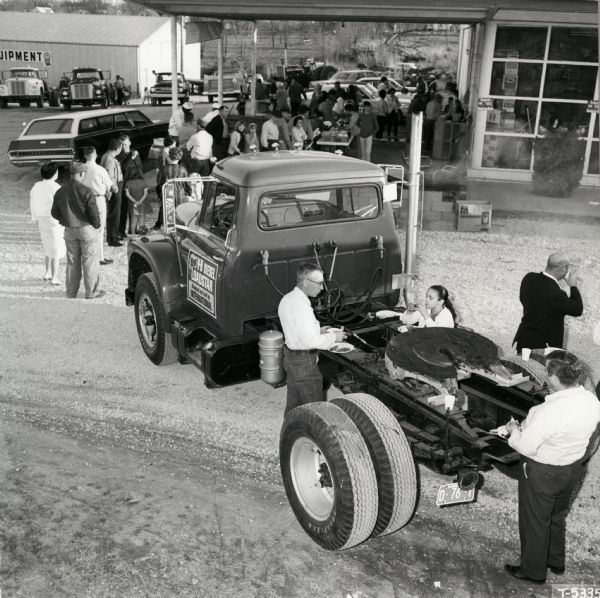 Elevated view of a crowd of farmers and businessmen gathered to attend International Harvester's "Diesel Demothon." In the foreground two men and a young girl are eating from paper plates from the bed of a truck.