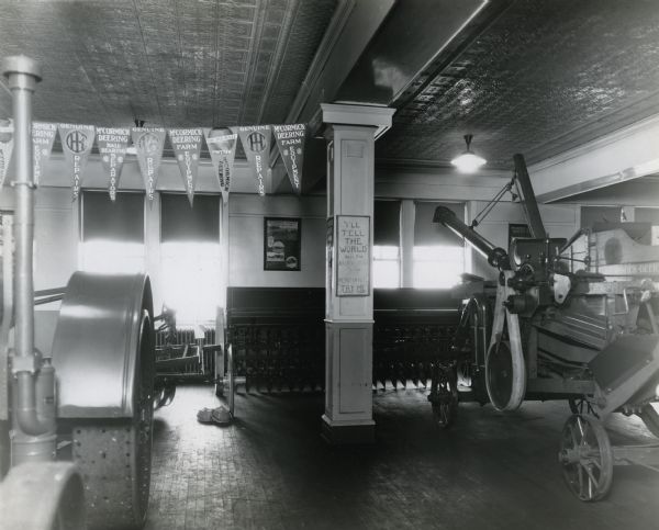 Farm machinery and equipment is arranged on the floor of International Harvester's Sioux City branch showroom. A string of pendants hangs from the ceiling, and a sign reading: "'I'll Tell the World' Says the Advertisement To The Merchant 'Try Me'" is posted to a pillar.