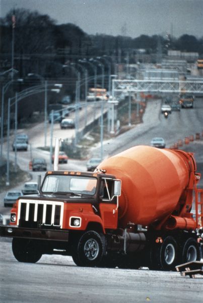 An International heavy S-2 cement mixer painted orange is at the top of a hill. In the background is a highway, part of which is under construction.