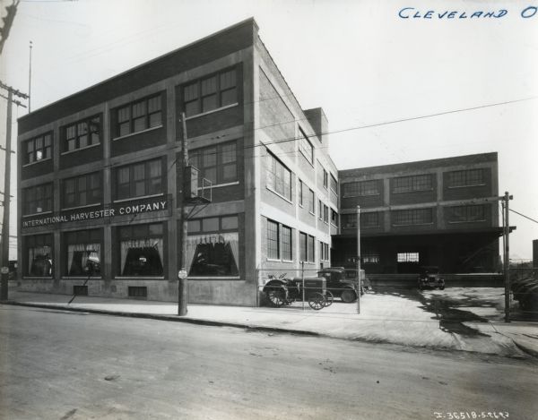 Exterior view of International Harvester Company's Cleveland branch building. Several automobiles and tractors are parked in a parking lot adjacent to a loading dock, and farm equipment is arranged on display in the first-floor showroom windows.
