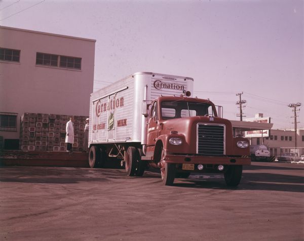 An International D-405 truck outfitted with a Trailmobile refrigerated trailer owned by Carnation Company backs up to a factory loading dock where two men are standing. The writing on the truck reads: "Carnation Fresh Ice Cream, Milk."