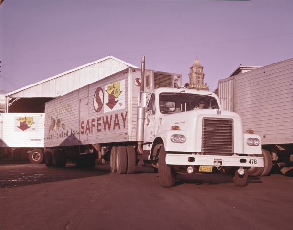An International model DT 405 truck outfitted with a Brown refrigerated trailer owned by Safeway Stores, Inc. pulls out from the loading dock of a building after delivering fresh milk. A tower with a sign reading "Safeway" is in the background.