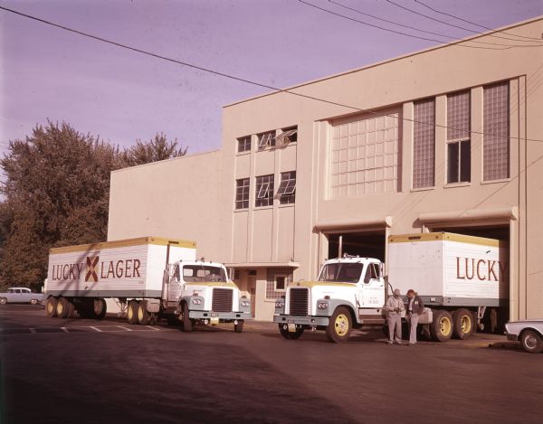 Two International DF-405 trucks with Fruehauf trailers owned by the Lucky Lager Corporation are parked in a loading area of a building. Two men stand near one of the trucks.