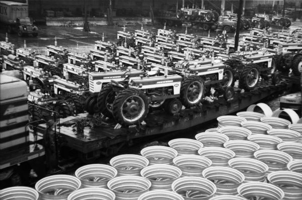Elevated view of Farmall tractors ready for delivery by rail and truck leave International Harvester's Farmall Works factory. In the foreground are stacks of what appears to be tire rims.