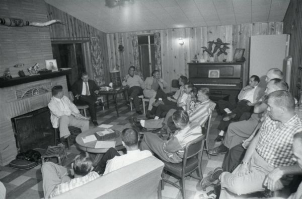 A group of men, probably International Harvester employees, sit in a room alongside a fireplace and piano during what appears to be a meeting.