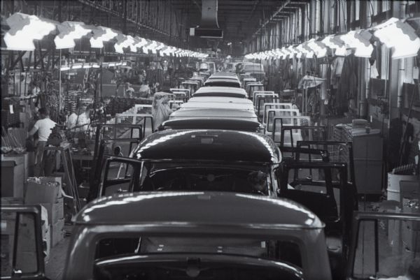 View over truck cabs on a long assembly line at International Harvester's Springfield Works. Factory workers are on both sides of the trucks.