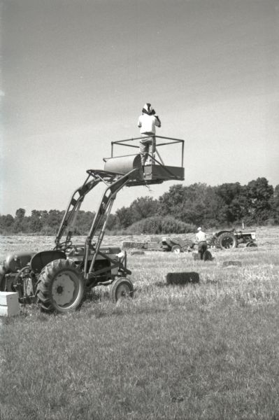 A man stands on an elevated platform attached to a tractor in a field while taking photographs of a hay baler in use at Hickory Hill, International Harvester's photo farm. The farm was located 75 miles southwest of Chicago and was kept as a location to photograph and make videos of International Harvester equipment.