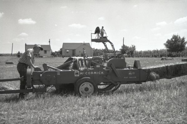 A man stands on a raised platform attached to a tractor with a camera while preparing to photograph a hay baler in a field on International Harvester's Hickory Hill farm. The farm was located 75 miles southwest of Chicago and was kept as a location to photograph and make videos of International Harvester equipment.