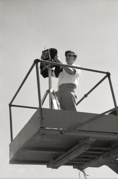 View from below of a male photographer standing on a platform with a camera while preparing to take photographs on International Harvester's Hickory Hill farm. The farm was located 75 miles southwest of Chicago and was kept as a location to photograph and make videos of International Harvester equipment.