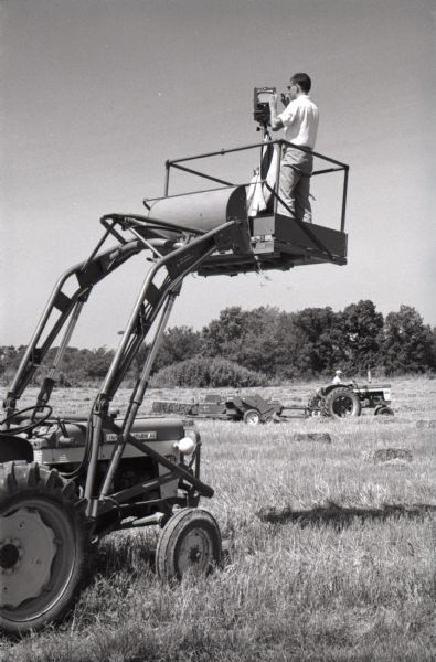 A man stands on a platform attached to an International tractor while taking photographs of a hay baler in a field at Hickory Hill. The farm was located 75 miles southwest of Chicago and was kept as a location to photograph and make videos of International Harvester equipment.