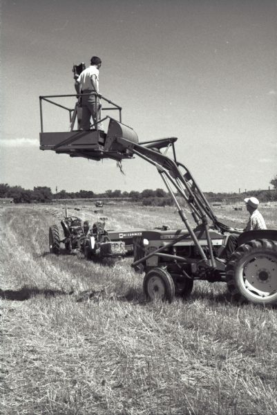 A photographer stands on a platform with his camera while taking photographs of a McCormick hay baler in use at Hickory Hill. The farm was located 75 miles southwest of Chicago and was kept as a location to photograph and make videos of International Harvester equipment.