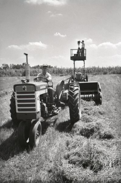 A male photographer stands on a platform with his camera while taking photographs of a McCormick hay baler in use at Hickory Hill. The farm was located 75 miles southwest of Chicago and was kept as a location to photograph and make videos of International Harvester equipment.