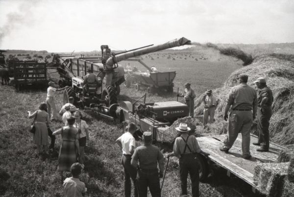 Elevated view of a men, women, and children gathered around a hay baler and other machines at work on International Harvester's Hickory Hill farm. The farm was located 75 miles southwest of Chicago and was kept as a location to photograph and make videos of International Harvester equipment.