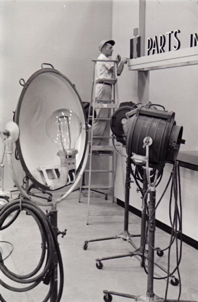A man stands on a ladder while installing what appears to be a backdrop in the studio on International Harvester's Hickory Hill farm. Several free-standing lights stand in the foreground. The farm was located 75 miles southwest of Chicago and was kept as a location to photograph and make videos of International Harvester equipment.