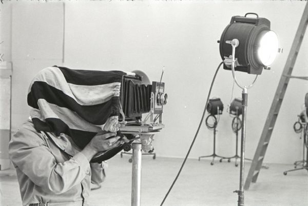 A man uses a cloth to cover his head as he looks through a view camera while photographing in the studio at Hickory Hill. The farm was located 75 miles southwest of Chicago and was kept as a location to photograph and make videos of International Harvester equipment.