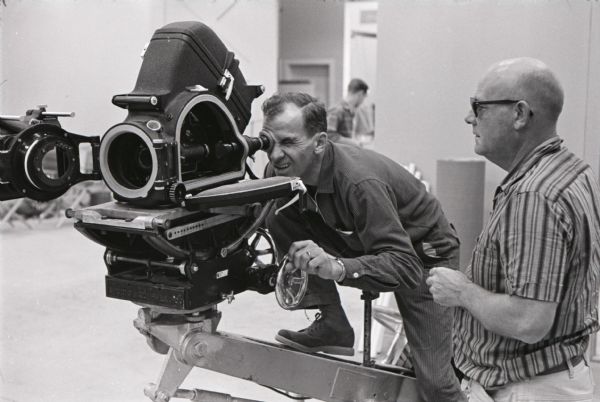 A man looks through a movie camera as another looks on in a studio at International Harvester's Hickory Hill Farm.