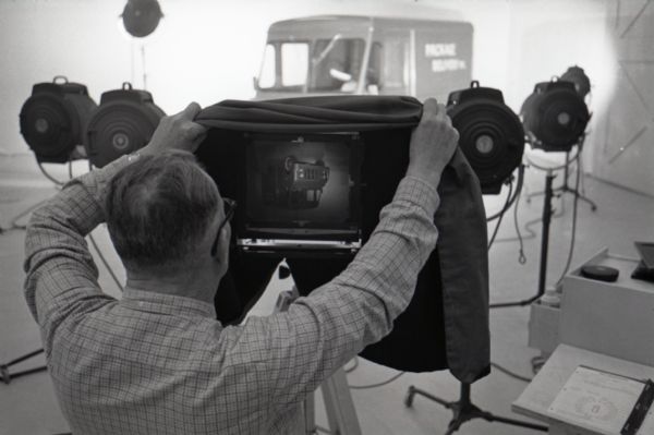 A man lifts the focusing cloth from the screen of a view camera while preparing to photograph an International Metro Mite truck inside a studio at International Harvester's Hickory Hill Farm.