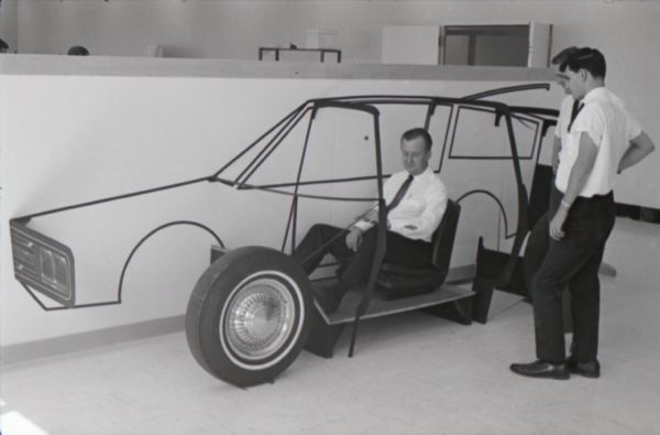 Men stand beside a model of a "Multipurpose Safety Utility Vehicle" while another man sits inside during the first styling seminar at the Fort Wayne motor truck engineering department. The program lasted ten weeks and participants were college students in their junior year.