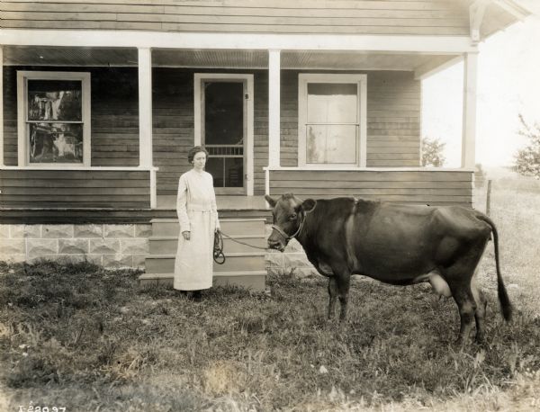 A woman holding a cow on a tether while standing in front of the porch in front of a farmhouse.