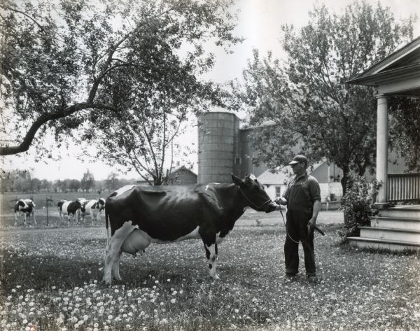 A man wearing a hat and overalls holding a dairy cow in front of a farmhouse. Trees, a barn with a silo, and other dairy cows are seen in the background. The original caption reads: "Earl Hughes R#1, Neenah, Wis."