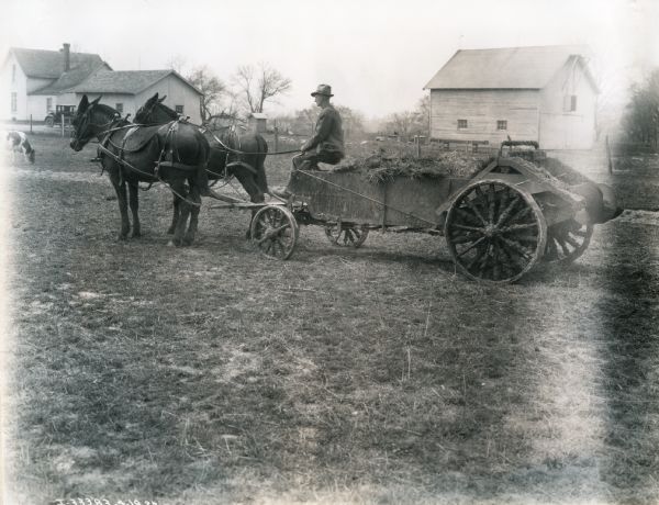 A man wearing a hat sitting on a two-horse manure spreader with farm buildings, an automobile, and a cow in the background. The original caption reads: "Kendall Stock Farm, Indianapolis, Ind."