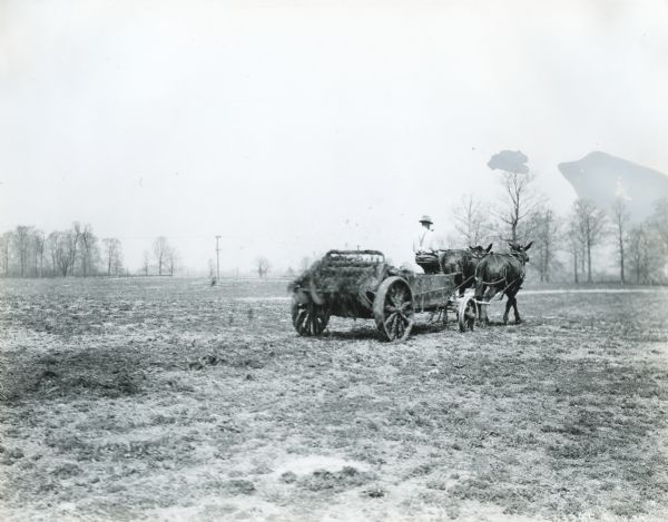 A man with a pipe in his mouth drives a team of horses to pull a manure spreader in a field. The original caption reads: "Kendall Stock Farm Indianapolis, Ind."